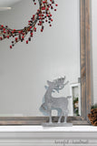 One paper reindeer decorations in front of a mirror under a red berry wreath.