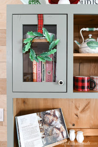 Gray hutch with natural wood interior and paper Christmas wreath hanging on the door.